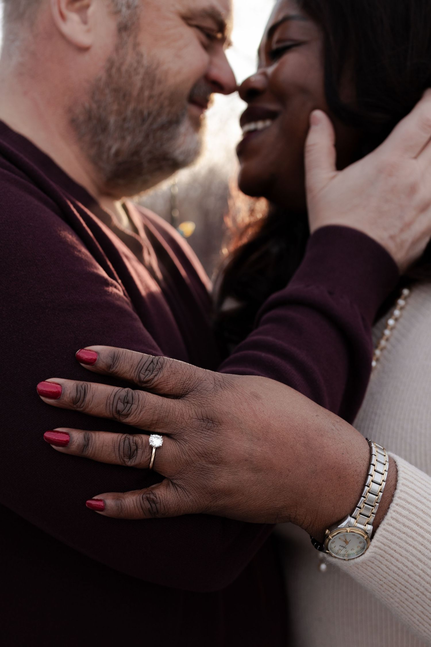 A joyful interracial couple embraces closely, the woman displaying an engagement ring and wearing a gold watch.