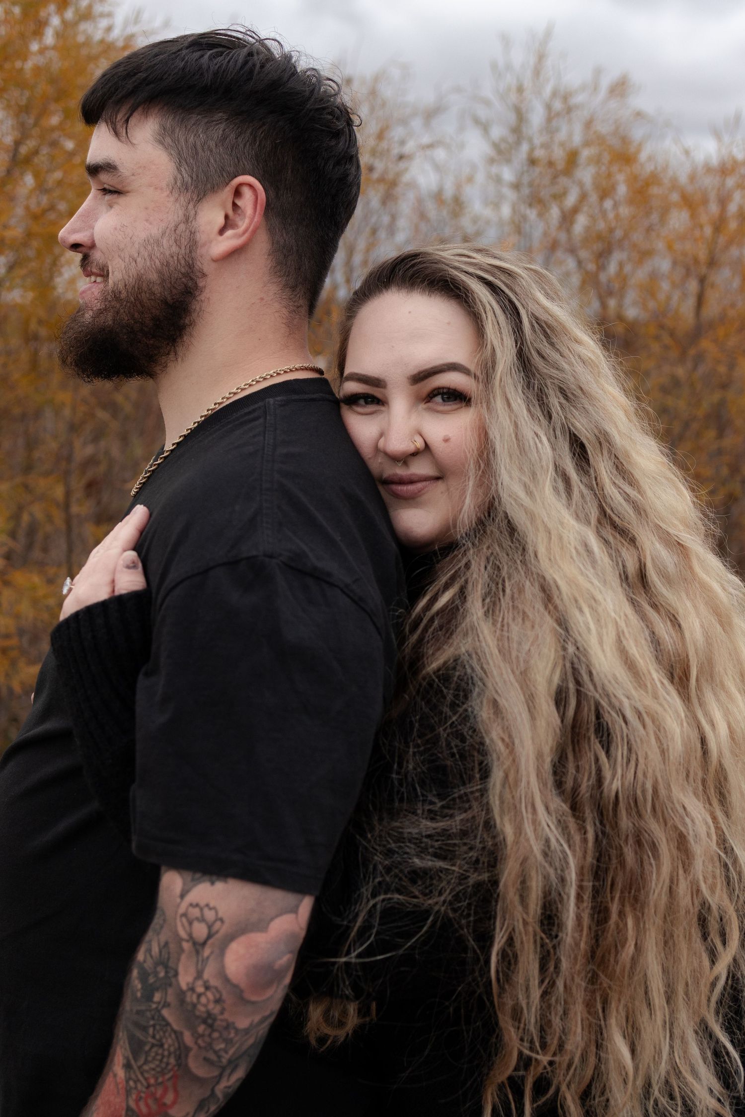 Woman with long blonde hair embraces man with arm tattoo from behind in autumn setting in Winnipeg