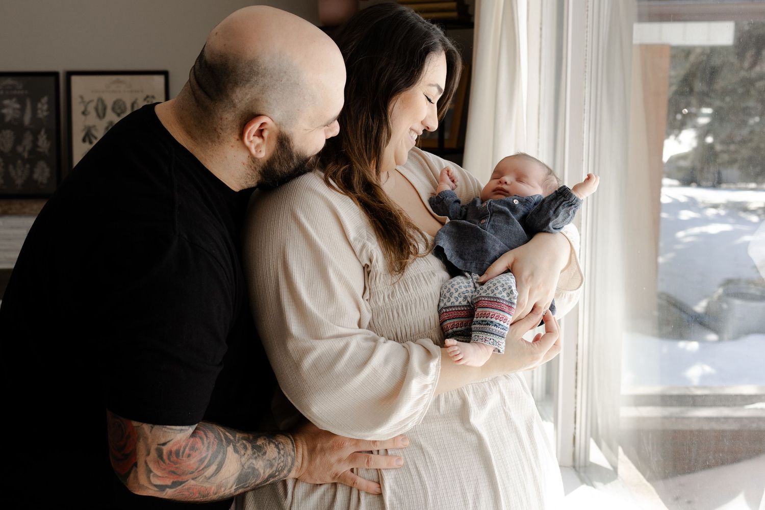 Couple with tattoos lovingly gazes at newborn baby by a snowy window in a warm family portrait.