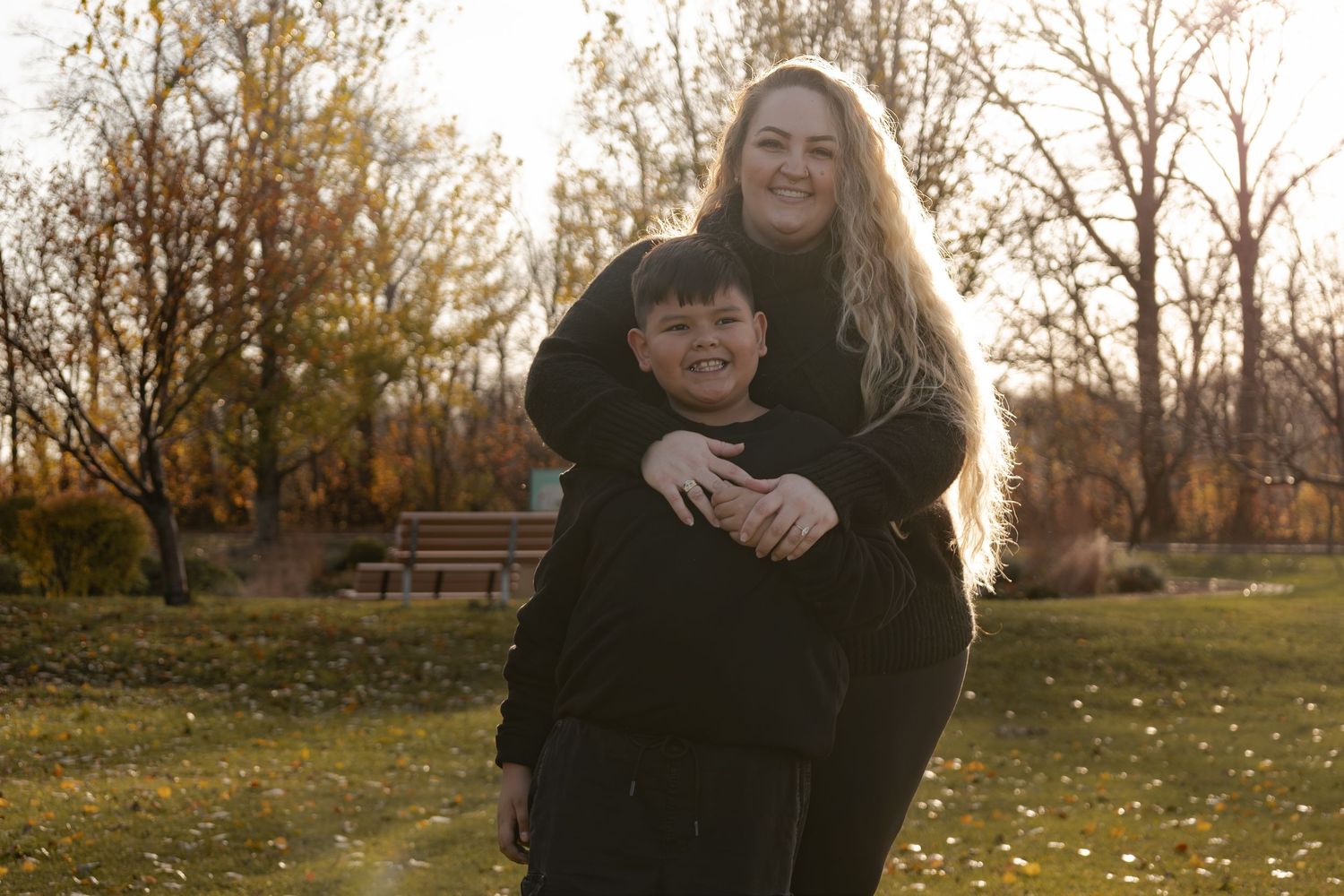 A woman hugs a boy from behind while smiling in a sunny autumn park.
