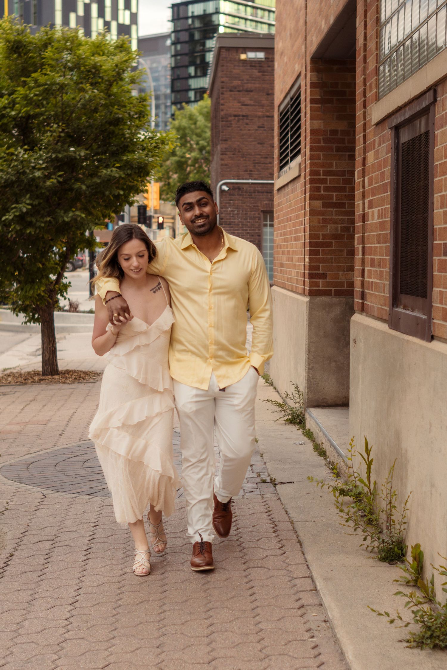 A happy interracial couple walks arm in arm on a city sidewalk, woman in cream dress, man in yellow shirt.