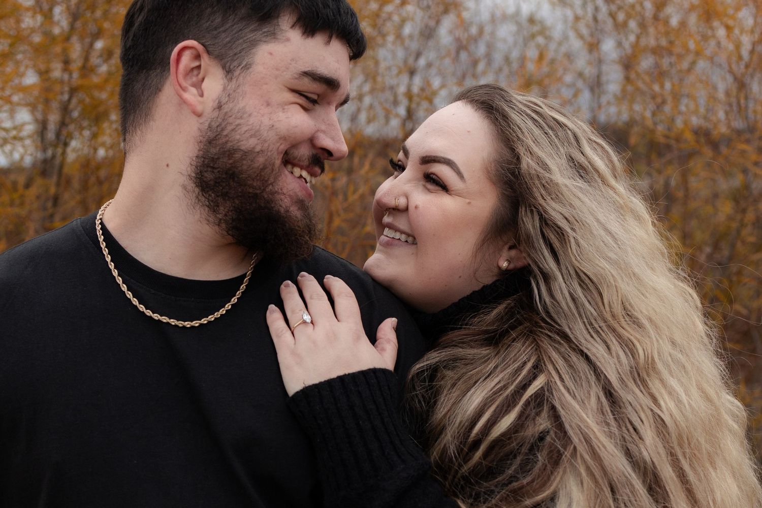 Happy couple smiling closely together outdoors in autumn, woman wearing engagement ring in warm fall setting in Winnipeg