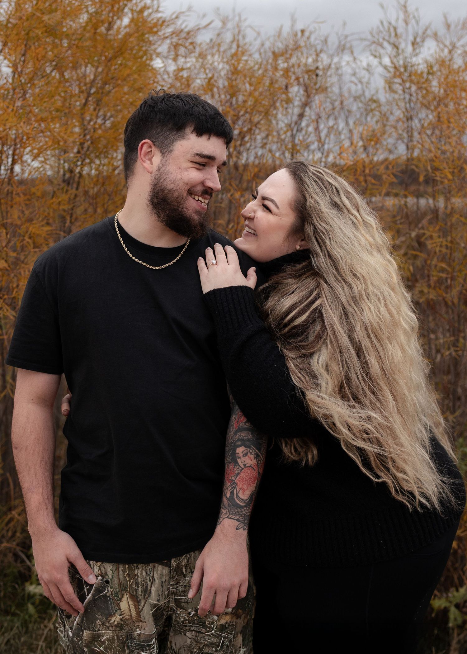 Smiling couple in black outfits standing together among autumn foliage, woman with tattoos resting hand on mans chest in Winnipeg