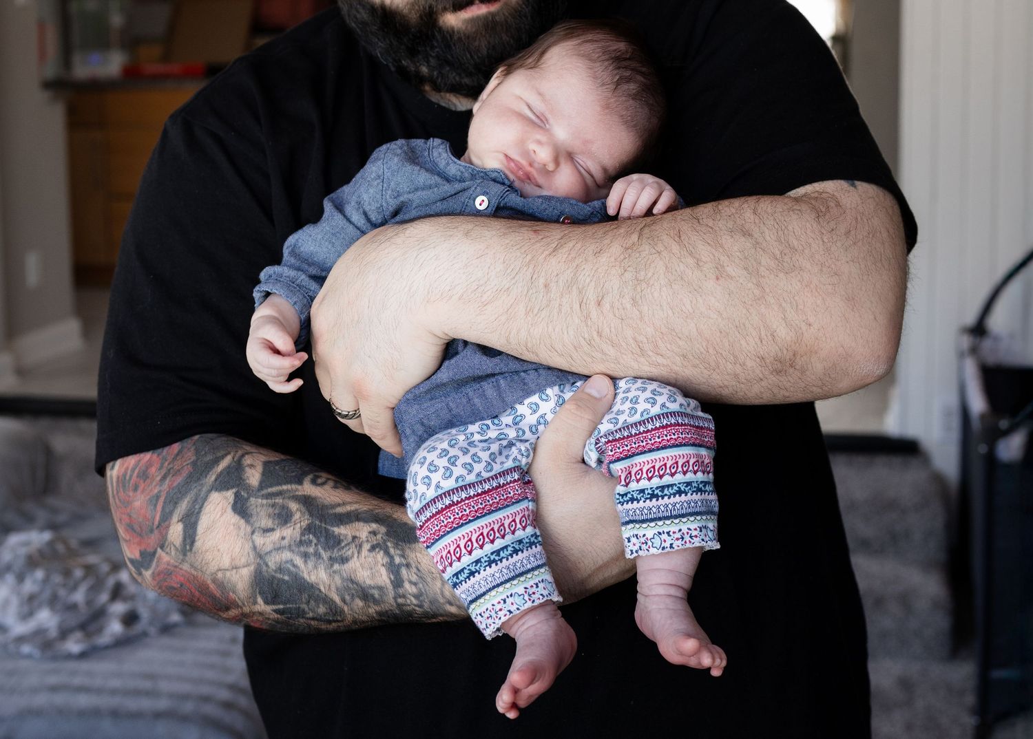 Tattooed father cradling sleeping newborn baby dressed in gray onesie and patterned pants.