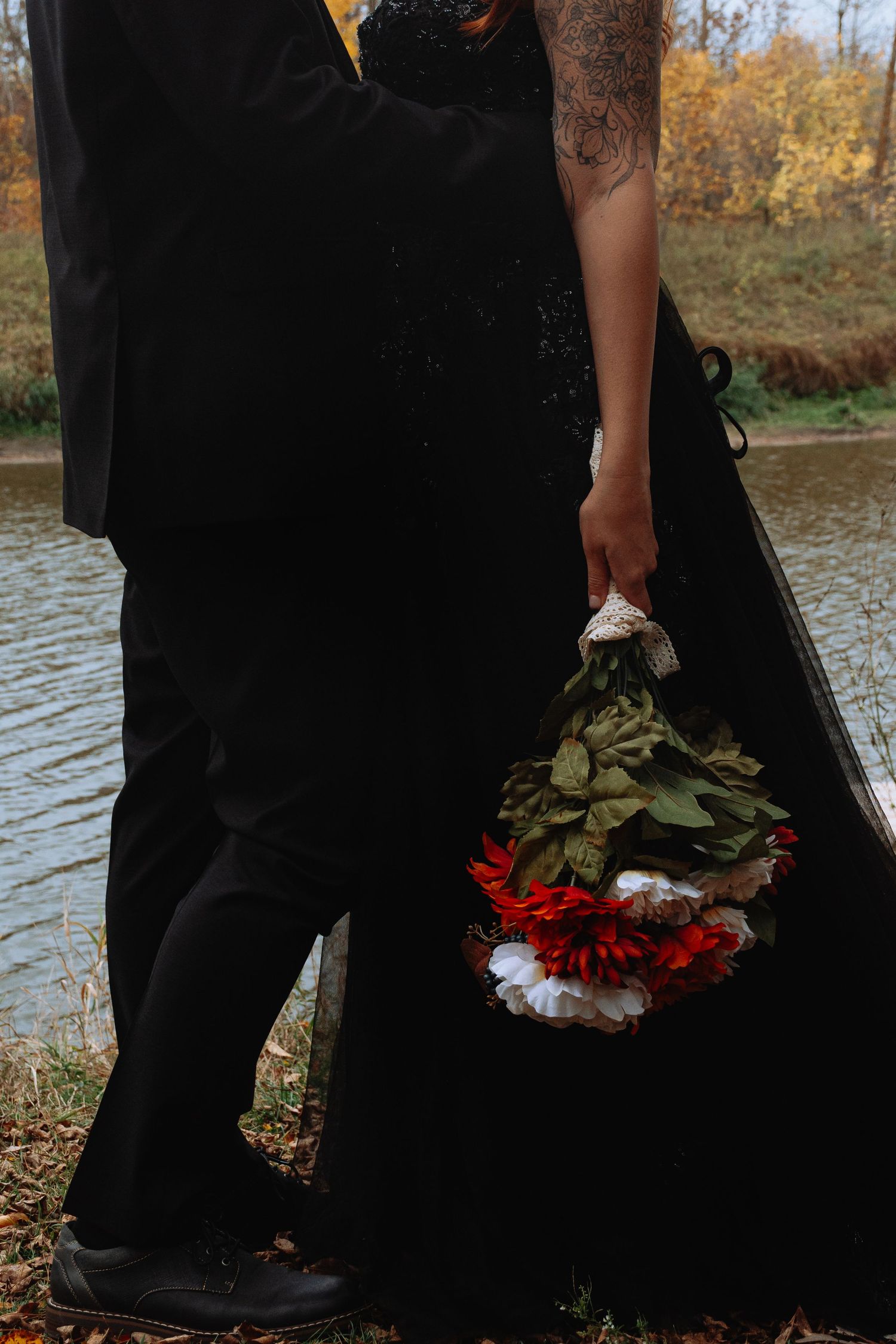 Couple in black wedding attire holding a red and white bouquet near an autumn lake.