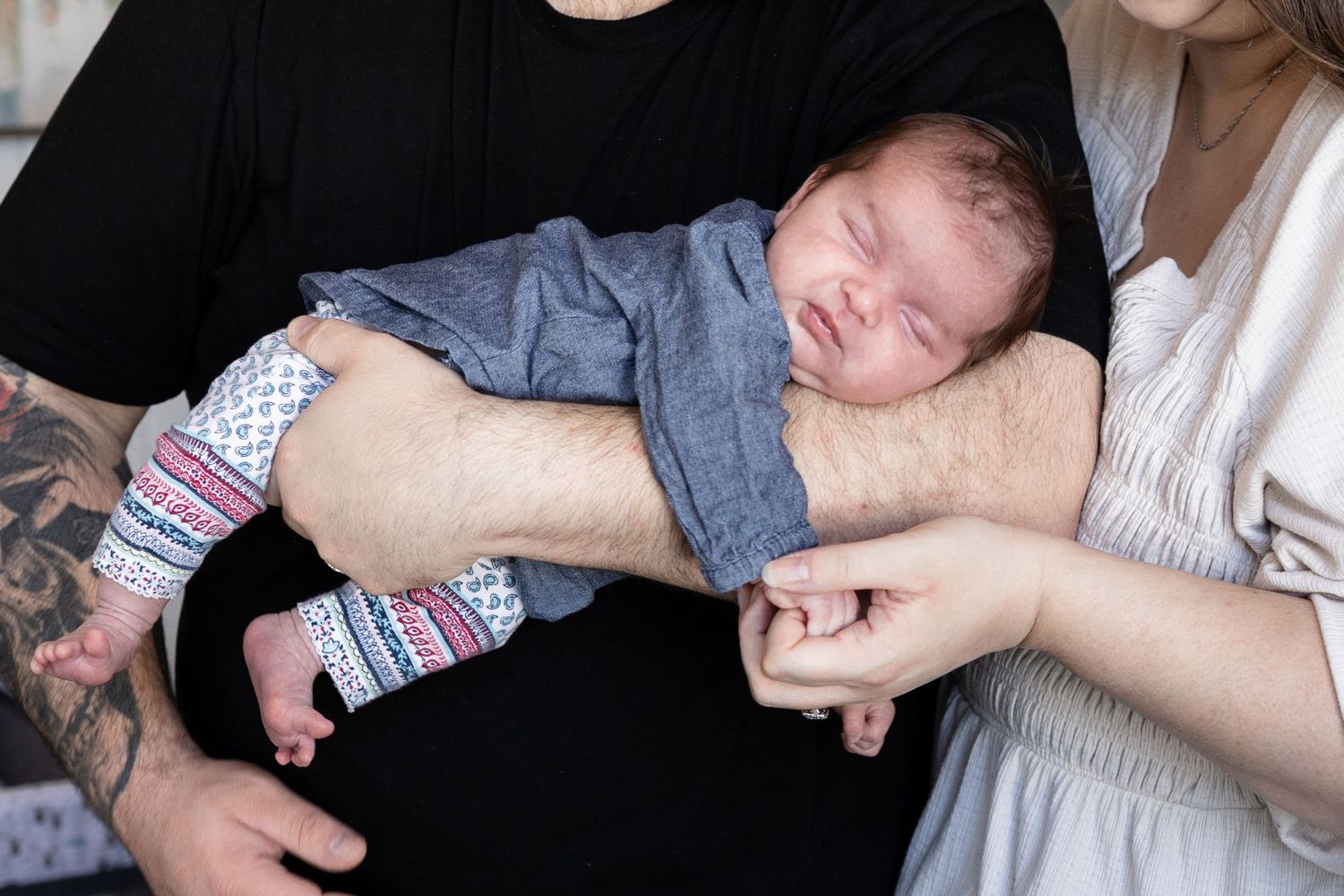 Newborn baby sleeping peacefully in father's tattooed arms while mother holds his tiny hand.