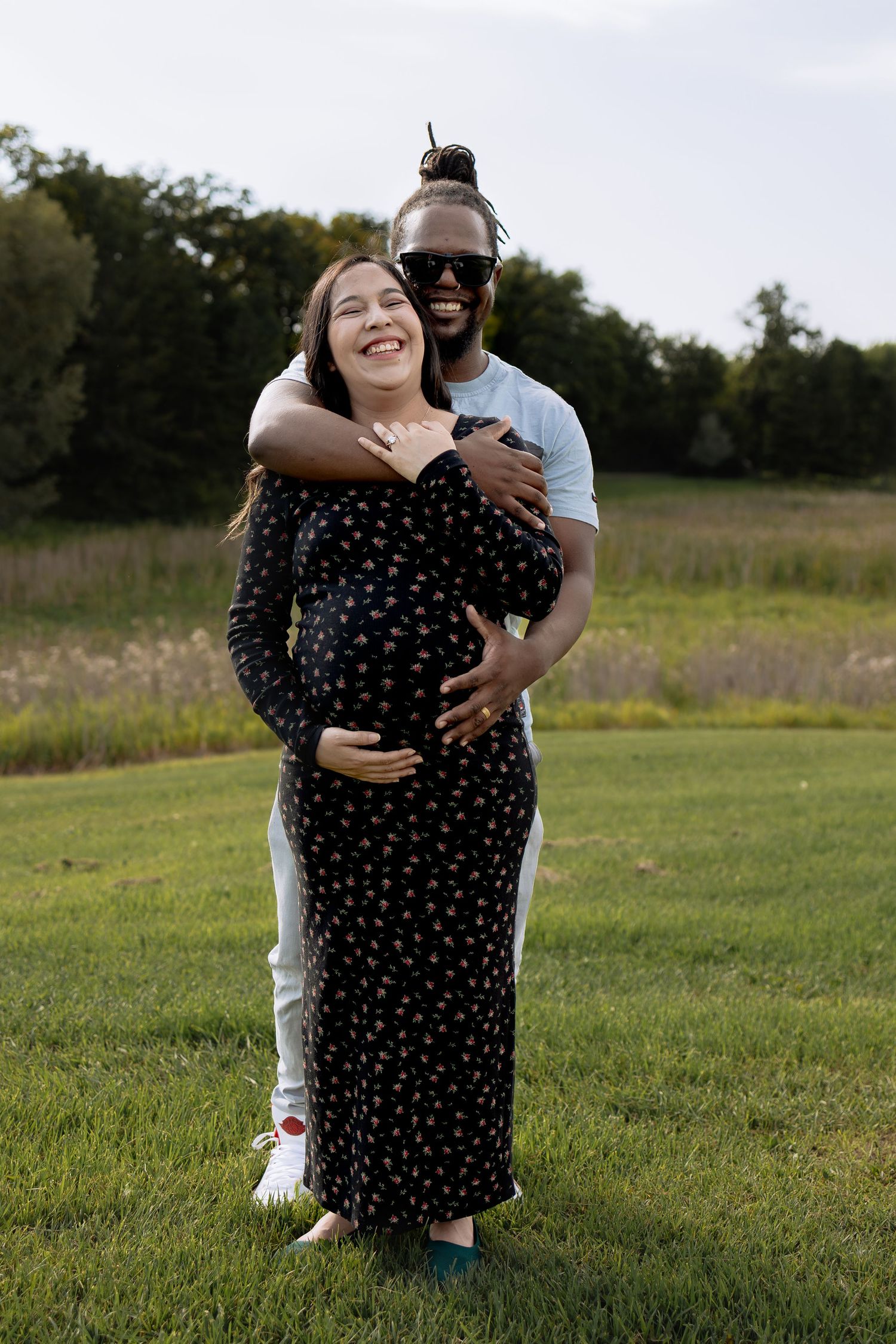 Interracial couple embracing outdoors, pregnant woman in black floral dress with partner hugging her belly in green field in Winnipeg