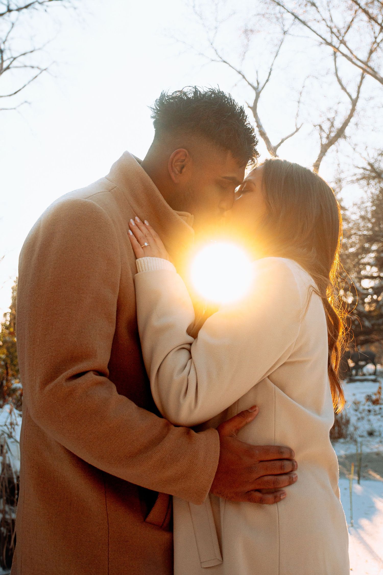 Couple sharing a romantic kiss outdoors in winter, backlit by golden sunlight with snowy trees behind them in Winnipeg