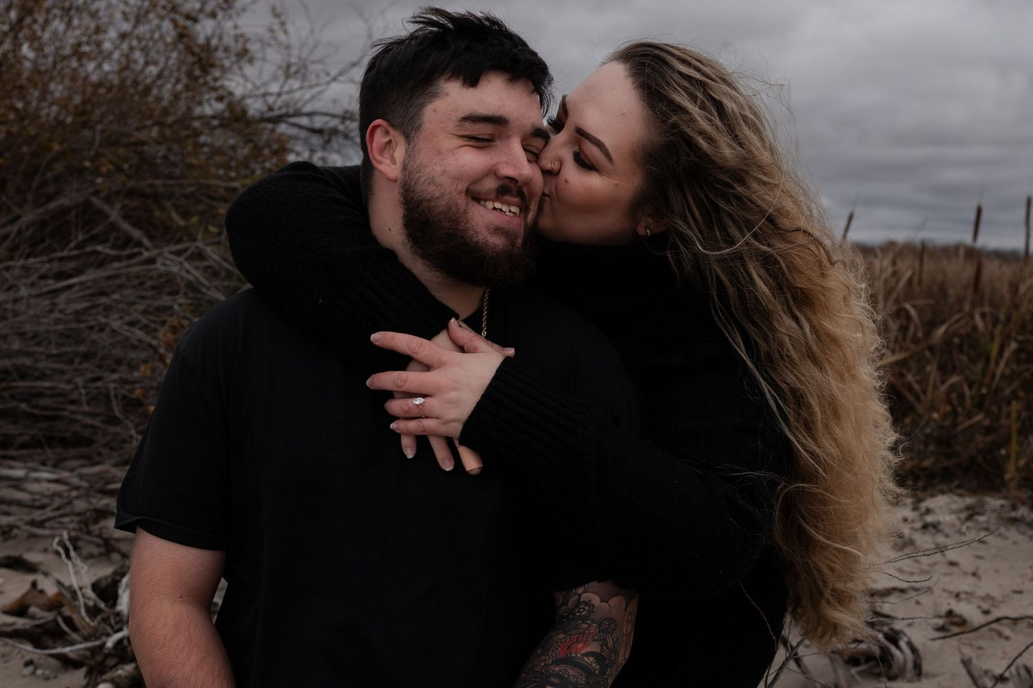 Happy couple embracing on sandy beach in matching black hoodies during fall engagement session in Winnipeg