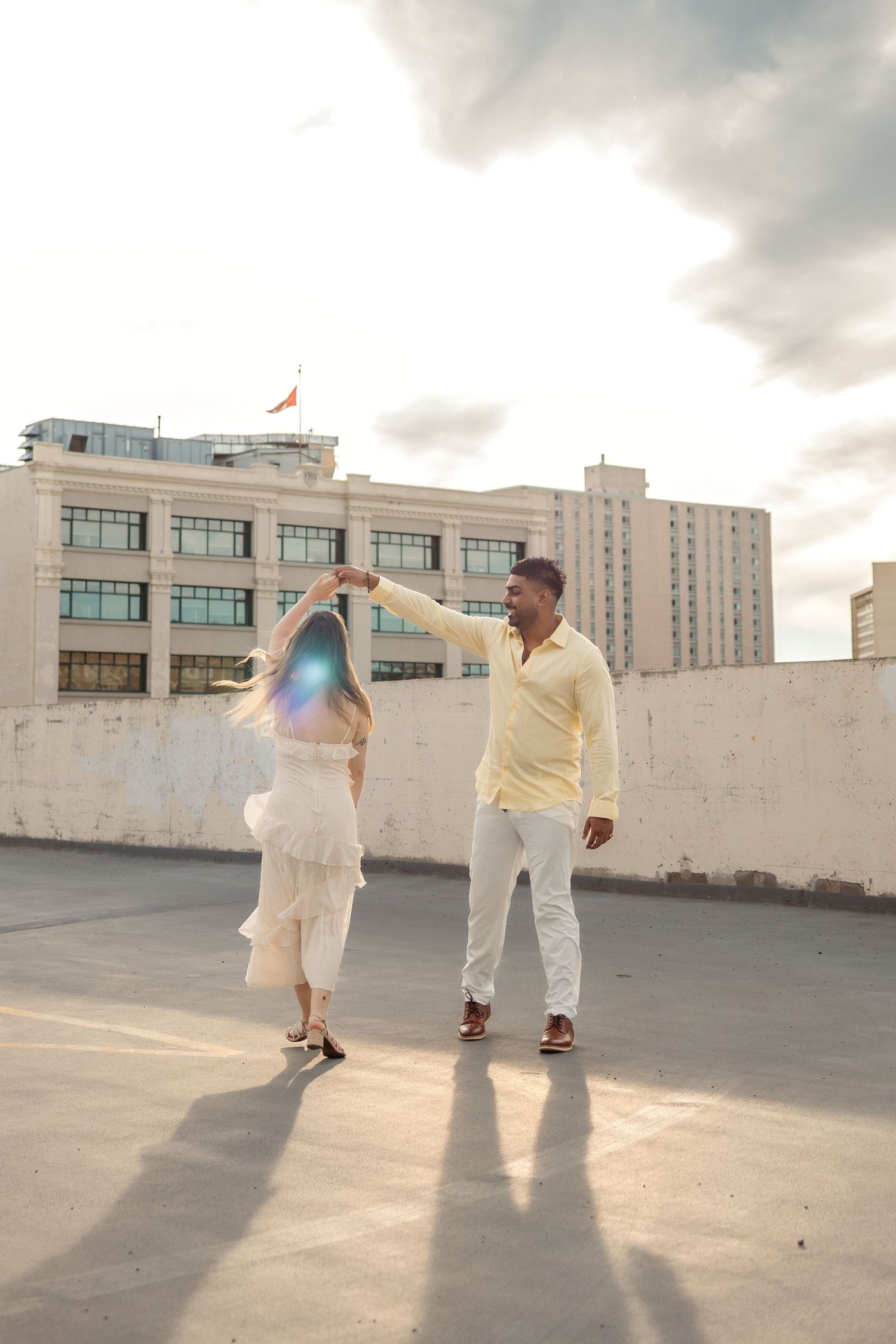 A couple dances joyfully on a rooftop, woman in white dress, man in yellow shirt, city buildings and cloudy sky behind them.