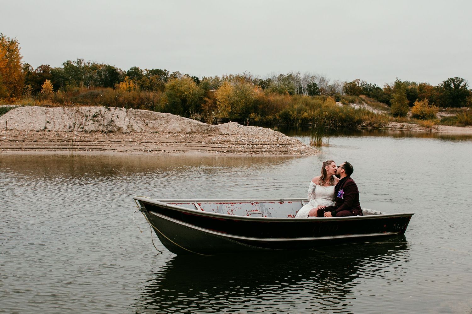 Bride and groom sharing a romantic kiss in a small boat on a calm autumn lake at a wedding in Winnipeg