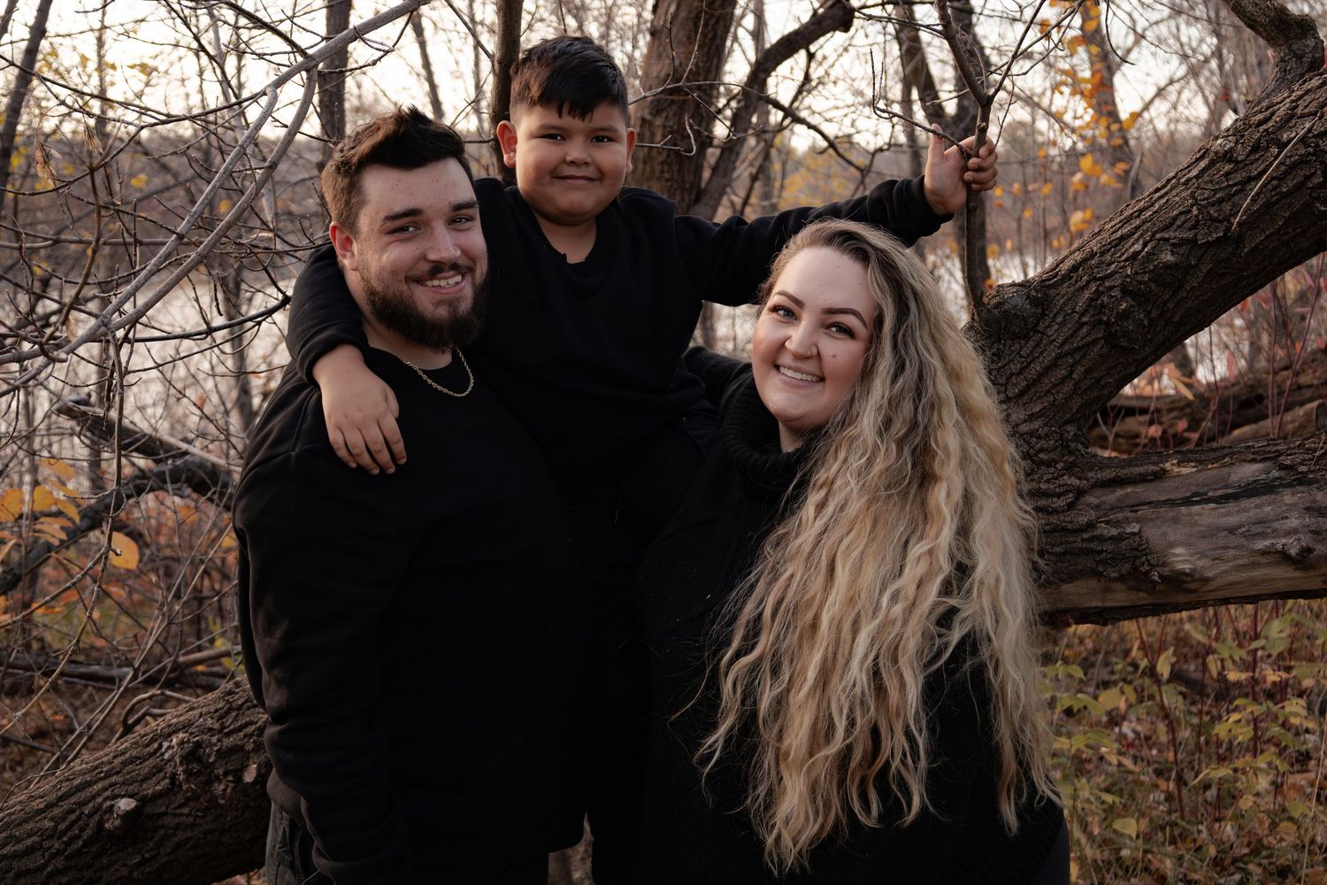 Smiling family of three posing playfully near a tree in an autumn woodland setting in Winnipeg