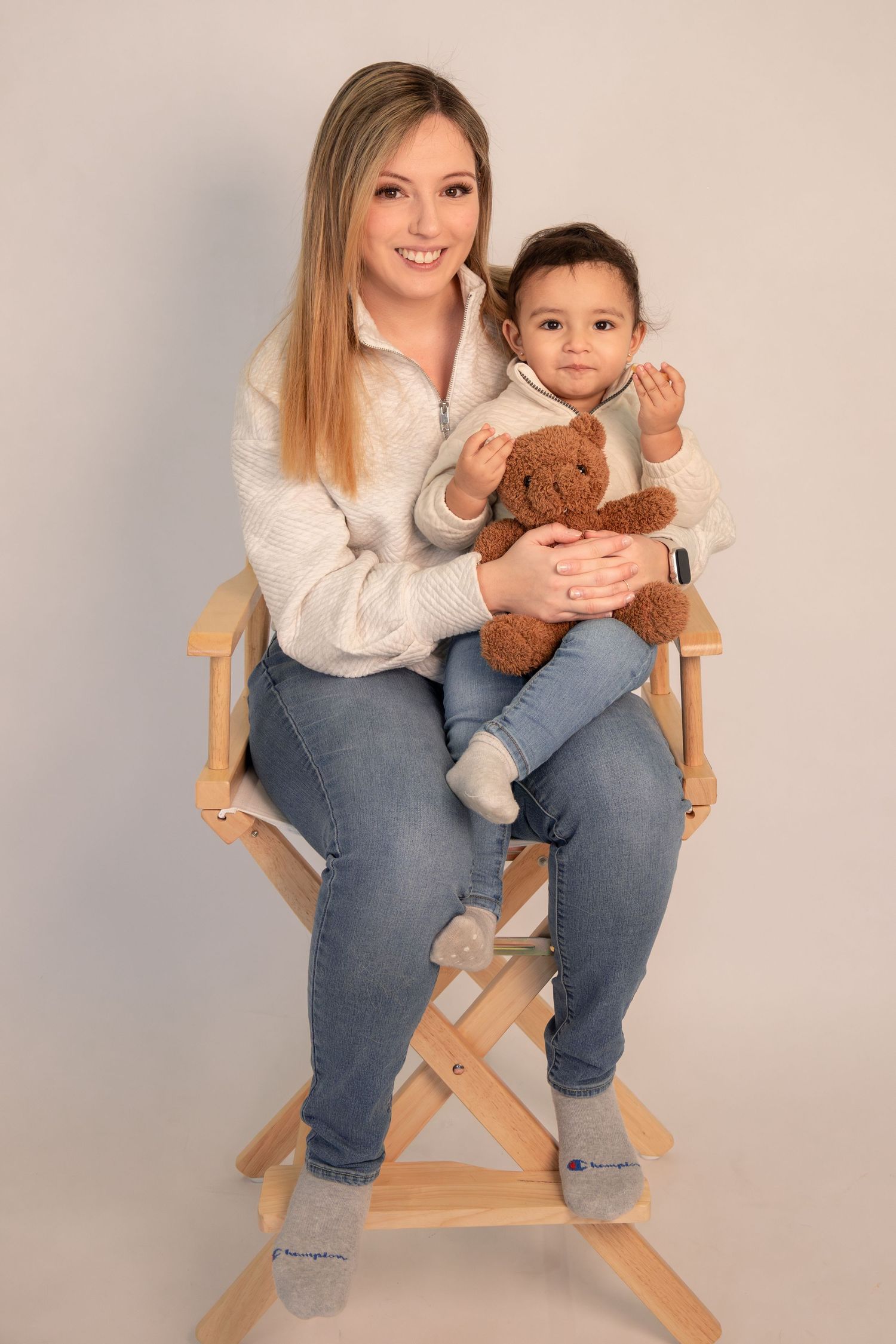 Smiling woman sitting on wooden chair holding a baby with a brown teddy bear in a studio setting.