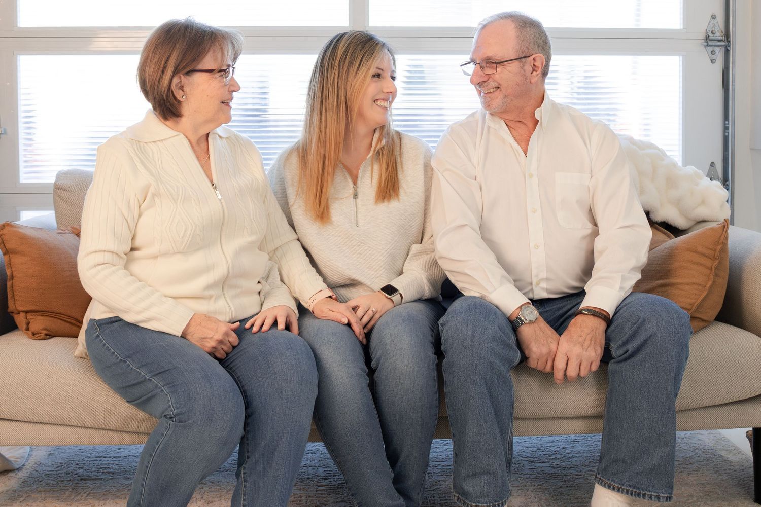 Three adults smiling and sitting together on a beige couch in a bright living room.
