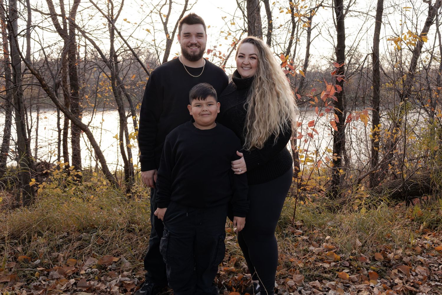 Family of three in matching black outfits posing outdoors among autumn foliage near a serene lake in Winnipeg