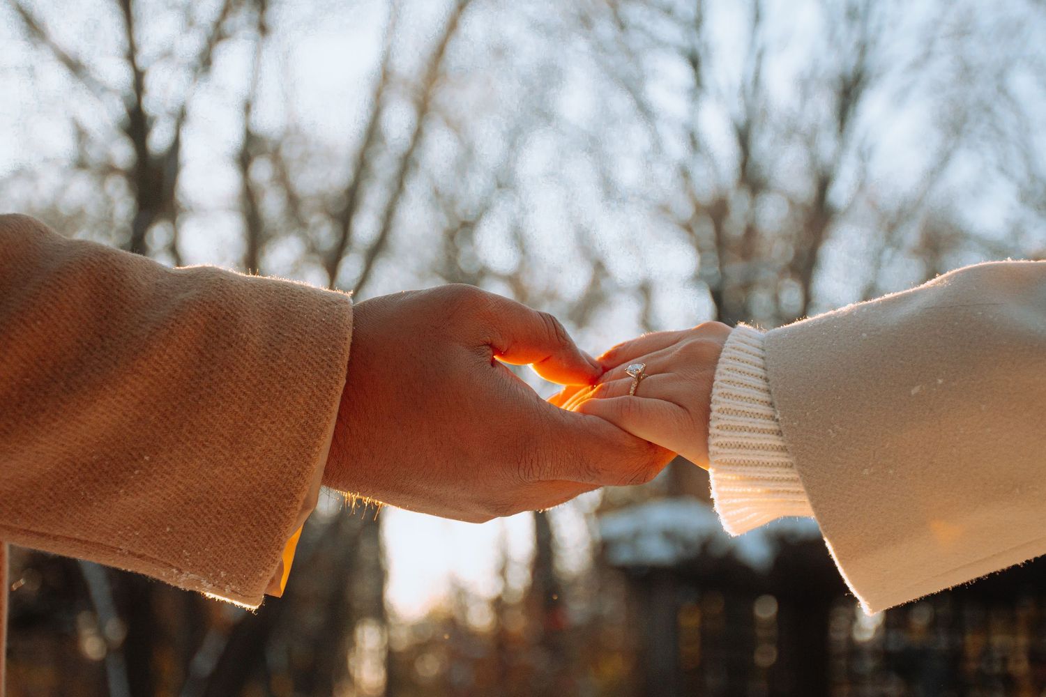 Close-up of couple in Winnipeg holding hands in warm coats with sunlit bokeh background outdoors.