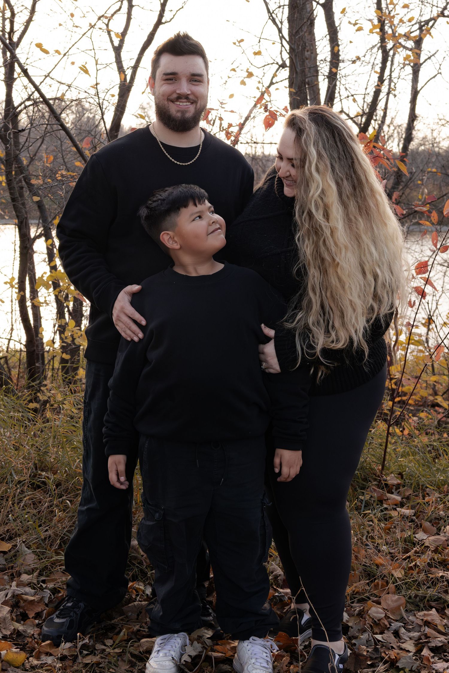Family of three in matching black outfits posing outdoors among autumn leaves near a river at sunset in Winnipeg