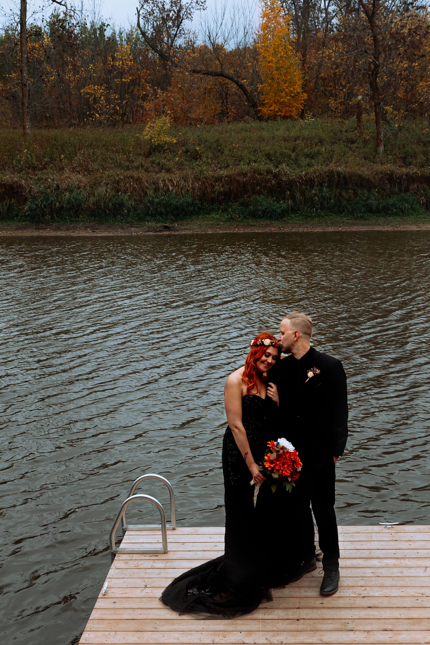 Bride and groom in matching black attire standing on a wooden dock beside an autumn lake.