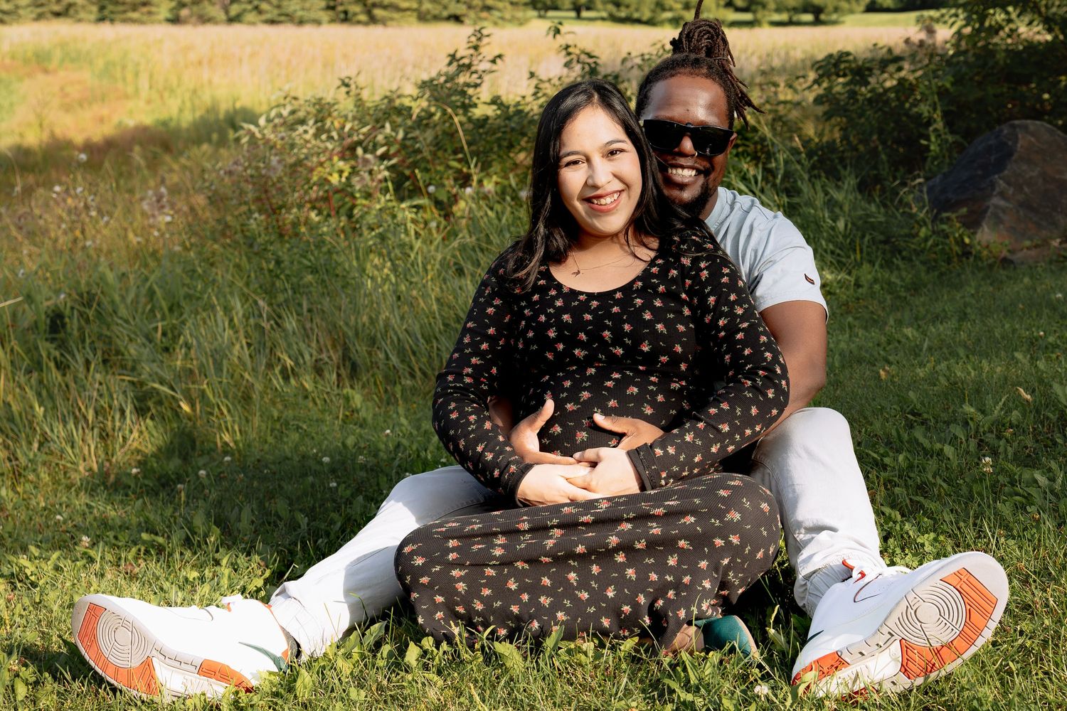 Happy interracial couple sitting together on grass in park cradling pregnant belly outdoors in Winnipeg