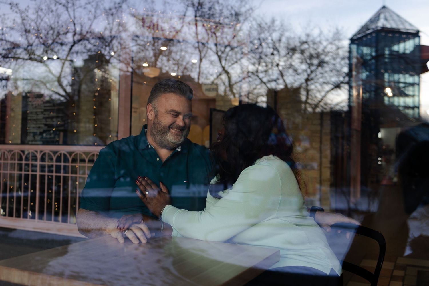 Two people smiling and talking at a table inside a cafe, viewed through a glass window at dusk.