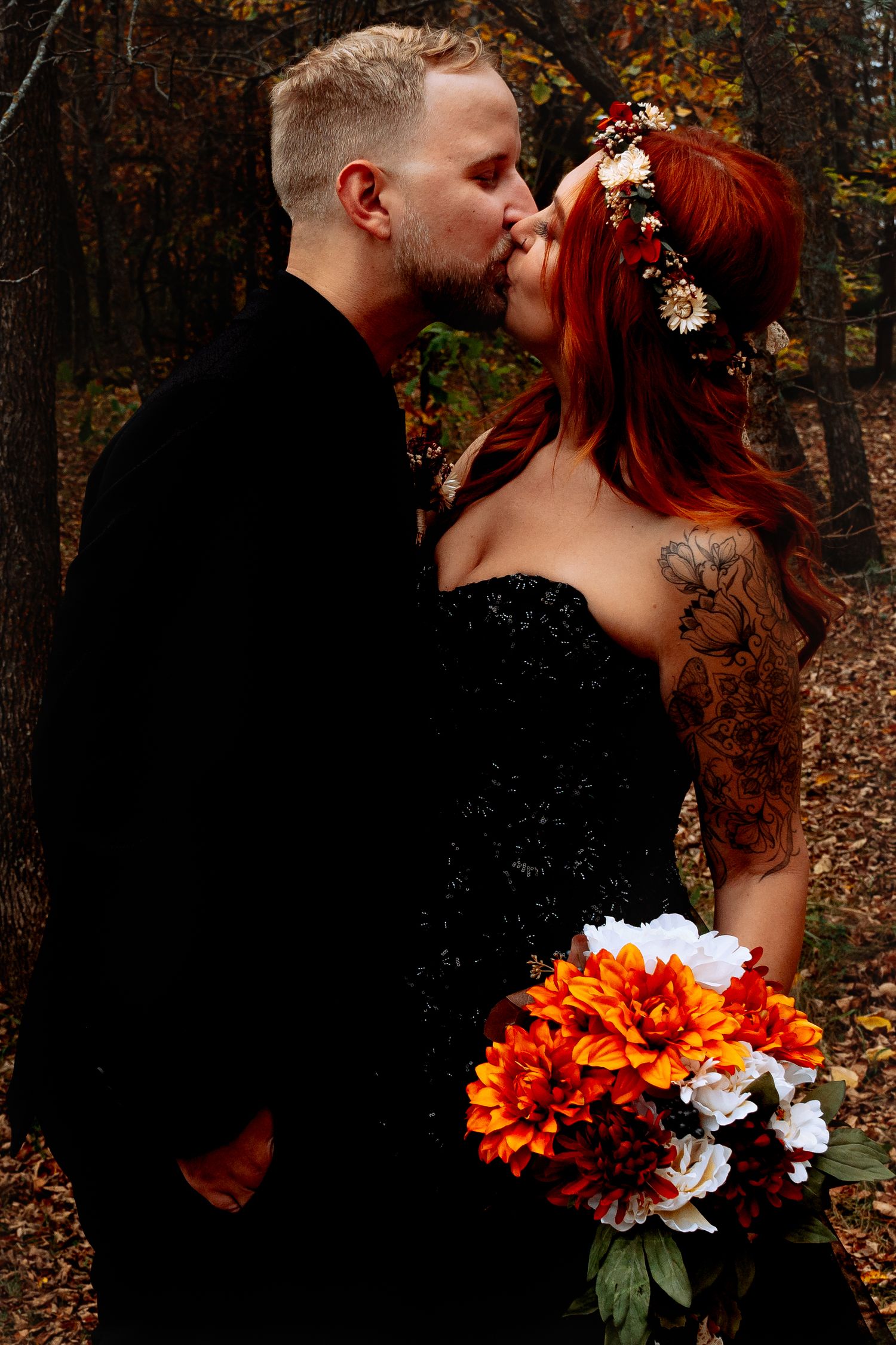 Groom kissing bride in black sequin dress with red hair and floral crown in autumn forest at a Wedding in Winnipeg