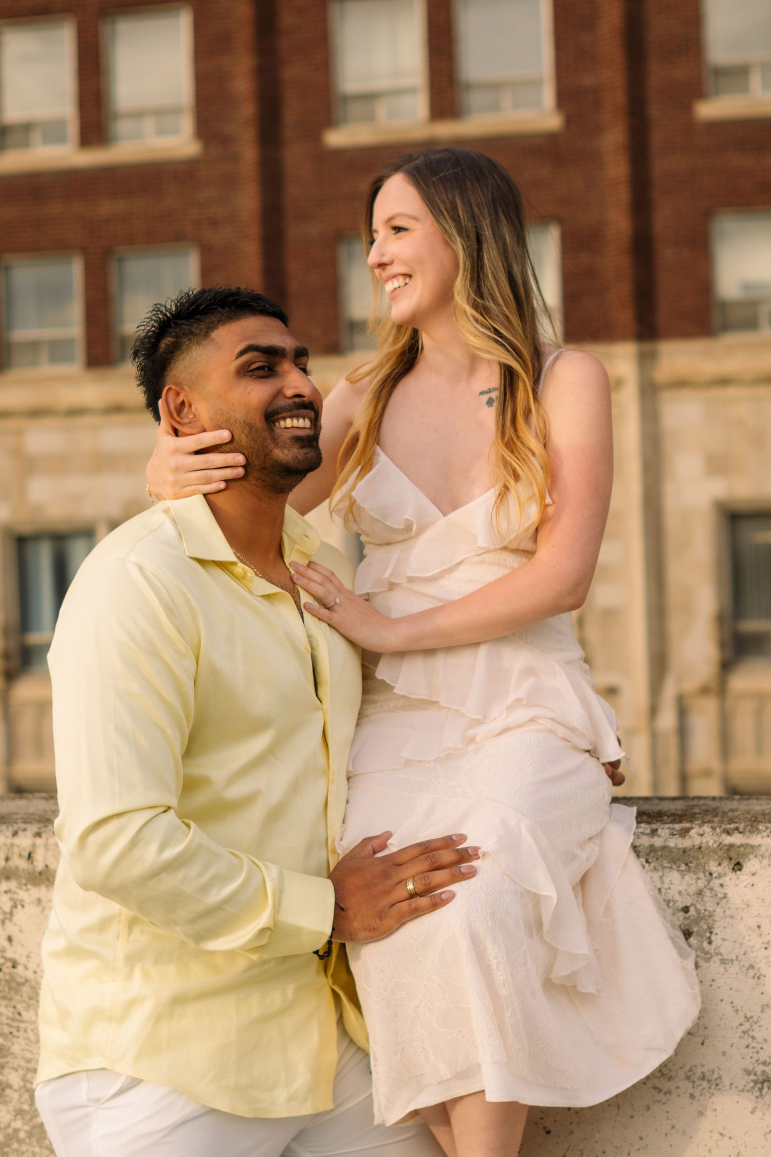A smiling couple in light-colored outfits share a joyful moment outdoors, with a brick building in the background.