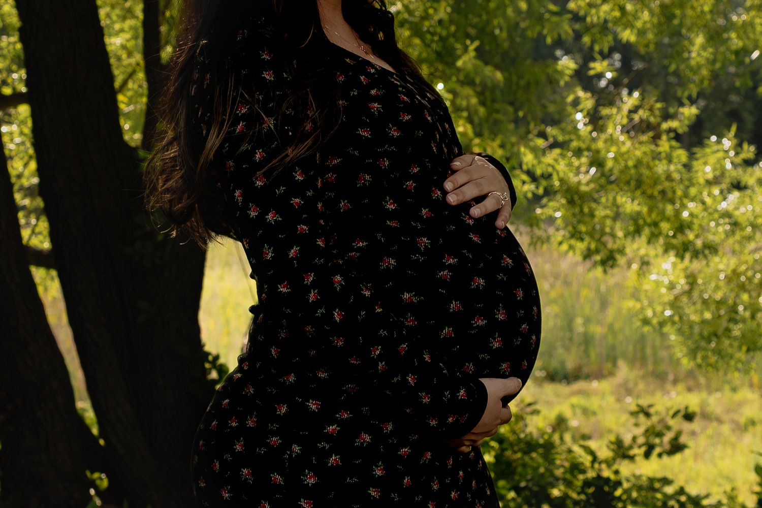 Pregnant woman in floral dress holding baby bump with trees in golden sunlight background in Winnipeg