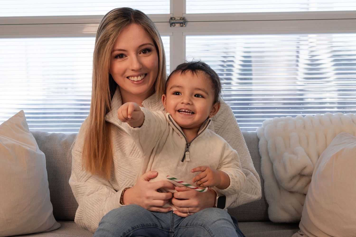 Young mother and toddler sitting on a gray sofa, smiling and pointing at the camera.