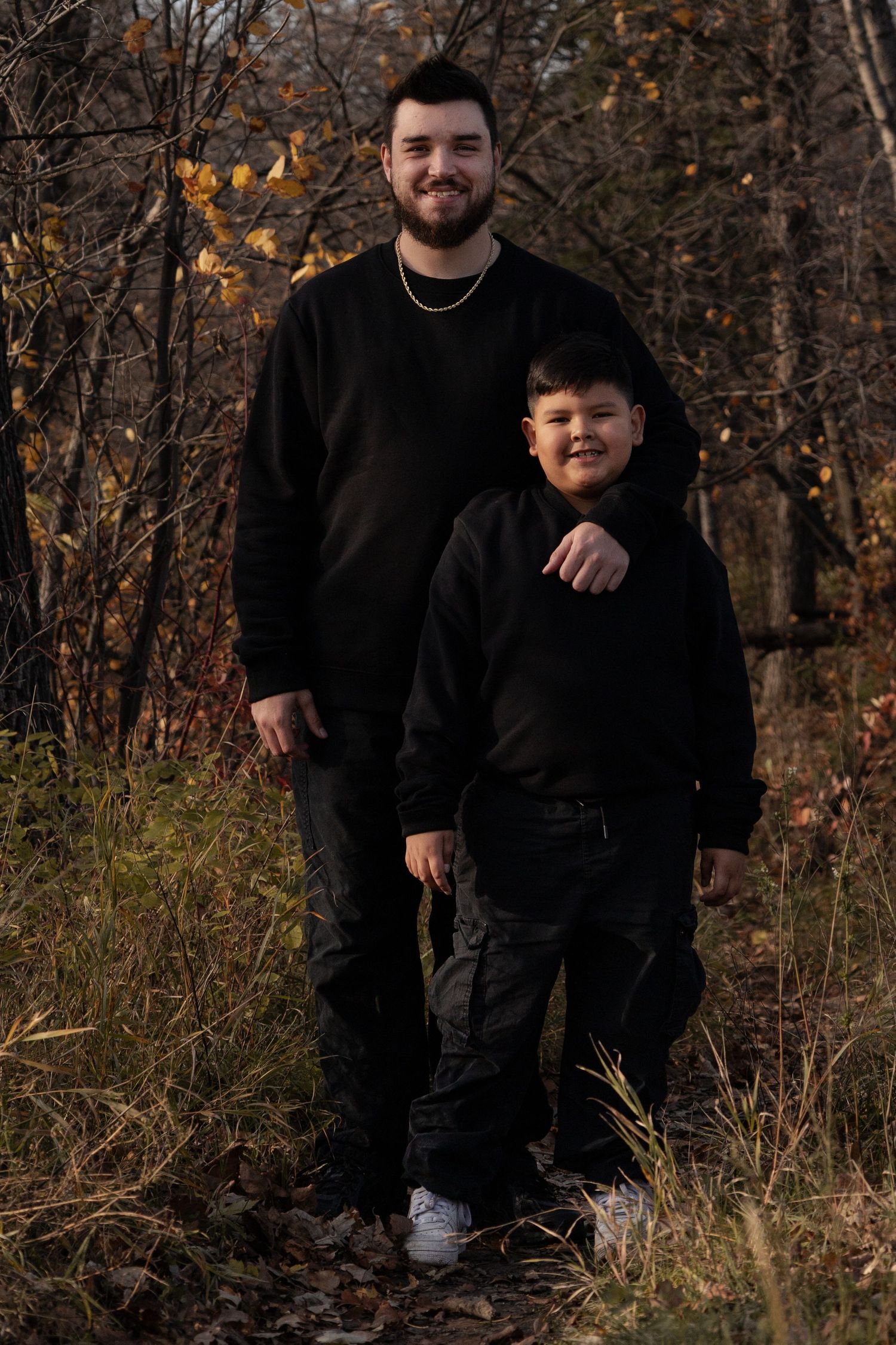 A young man and boy in matching black outfits pose in an autumn forest setting in Winnipeg