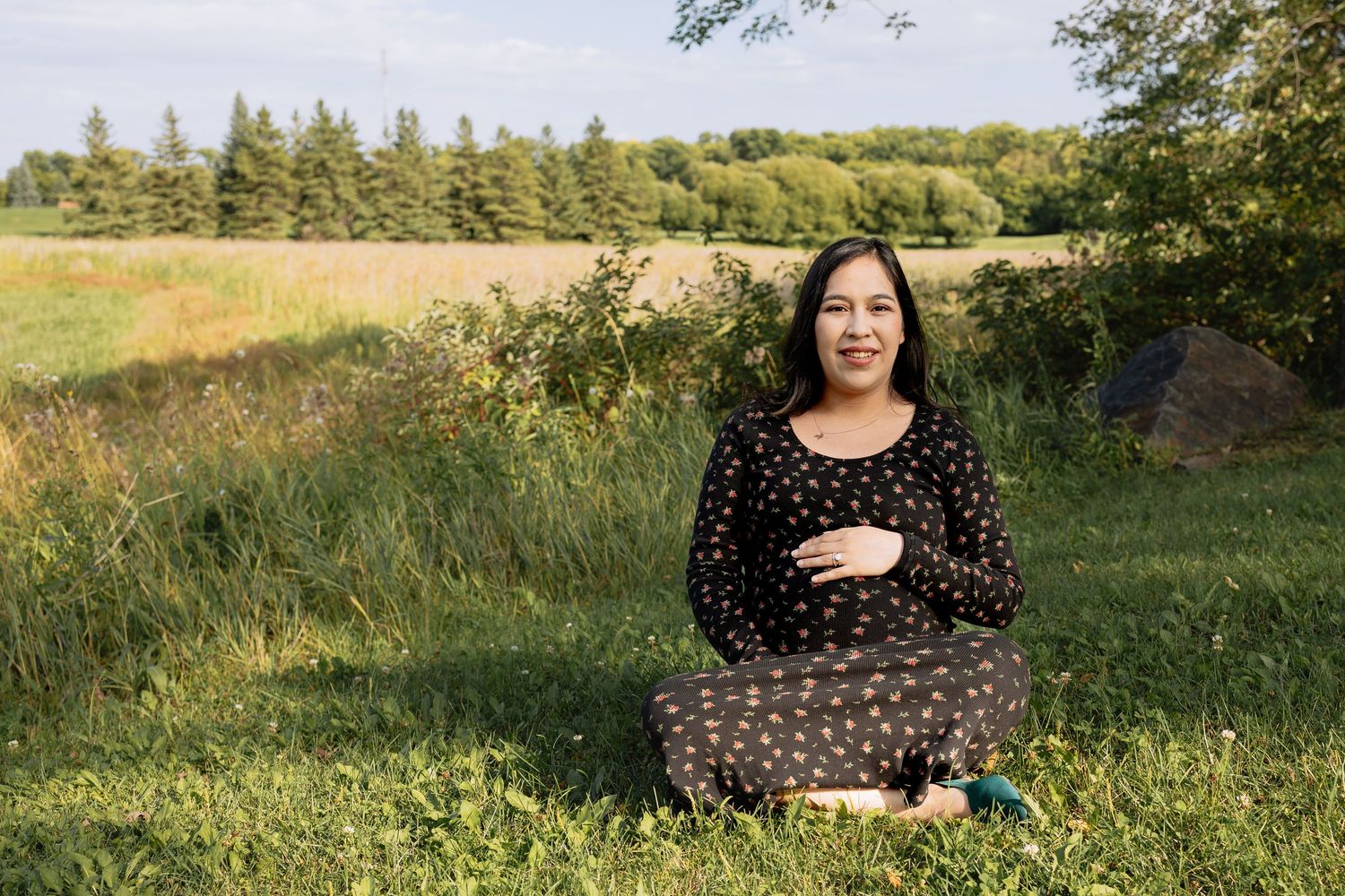 Smiling pregnant woman sitting on green grass in countryside wearing floral maternity dress in Winnipeg