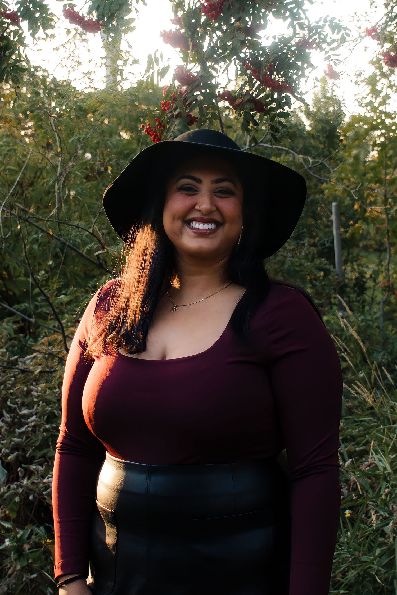 Smiling woman in burgundy top and black hat posing outdoors with red berries and greenery behind her in Winnipeg