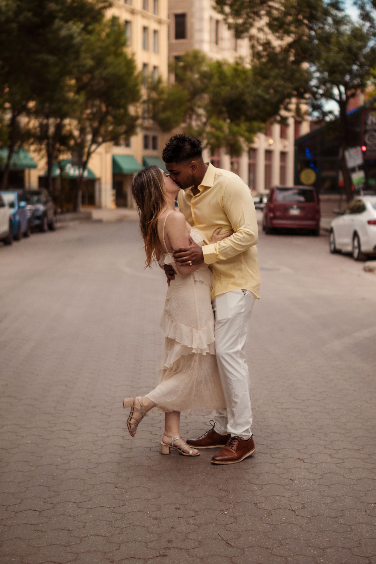 An interracial couple shares a romantic kiss in the middle of a cobblestone city street, dressed in elegant cream and yellow attire.