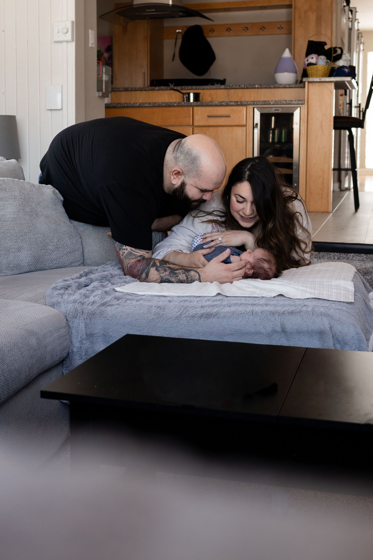 A bald tattooed father leans over a gray sofa to kiss his smiling wife holding their newborn baby at home.