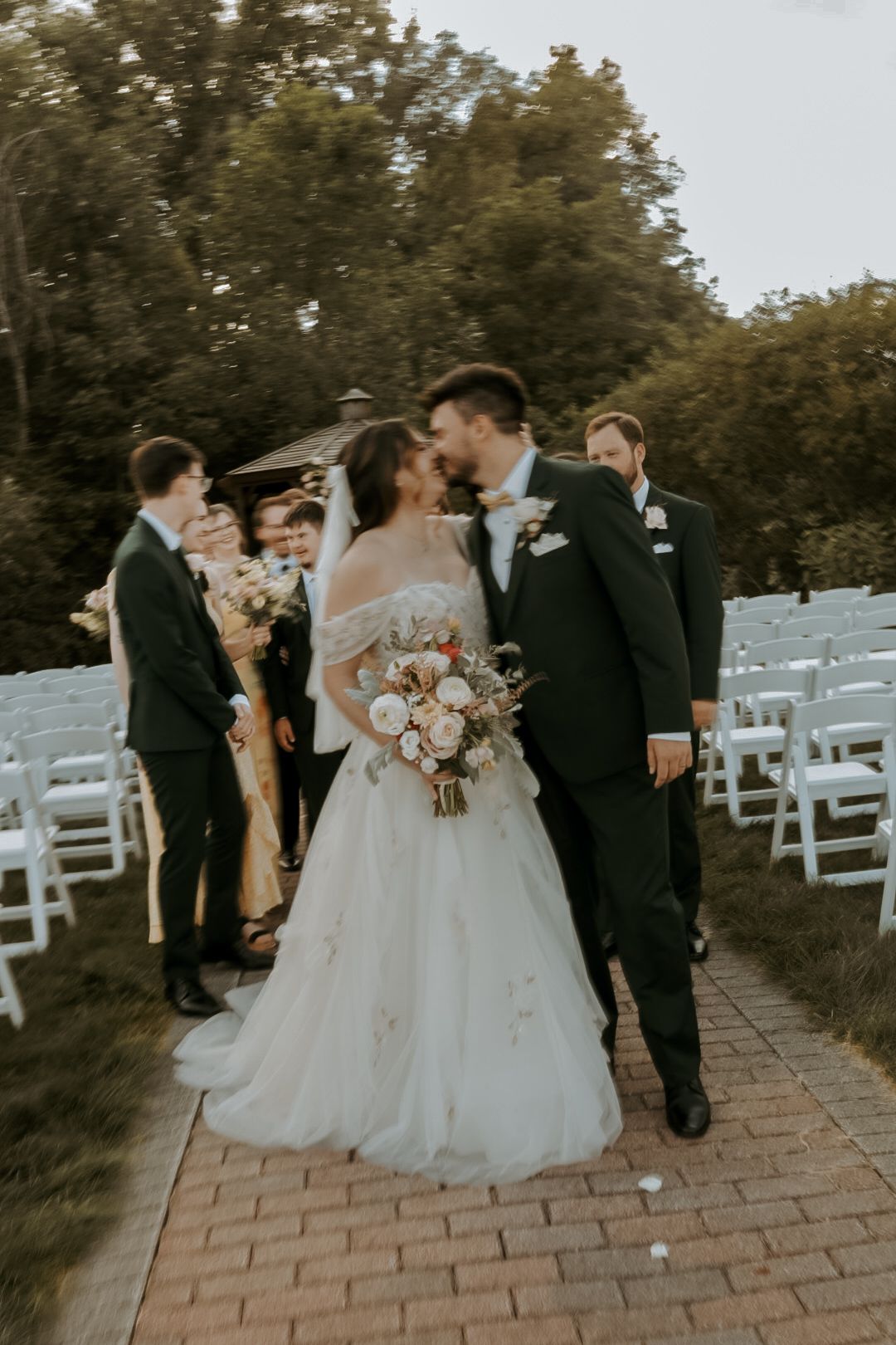Wedding party celebrates on a brick pathway surrounded by white chairs and lush greenery.