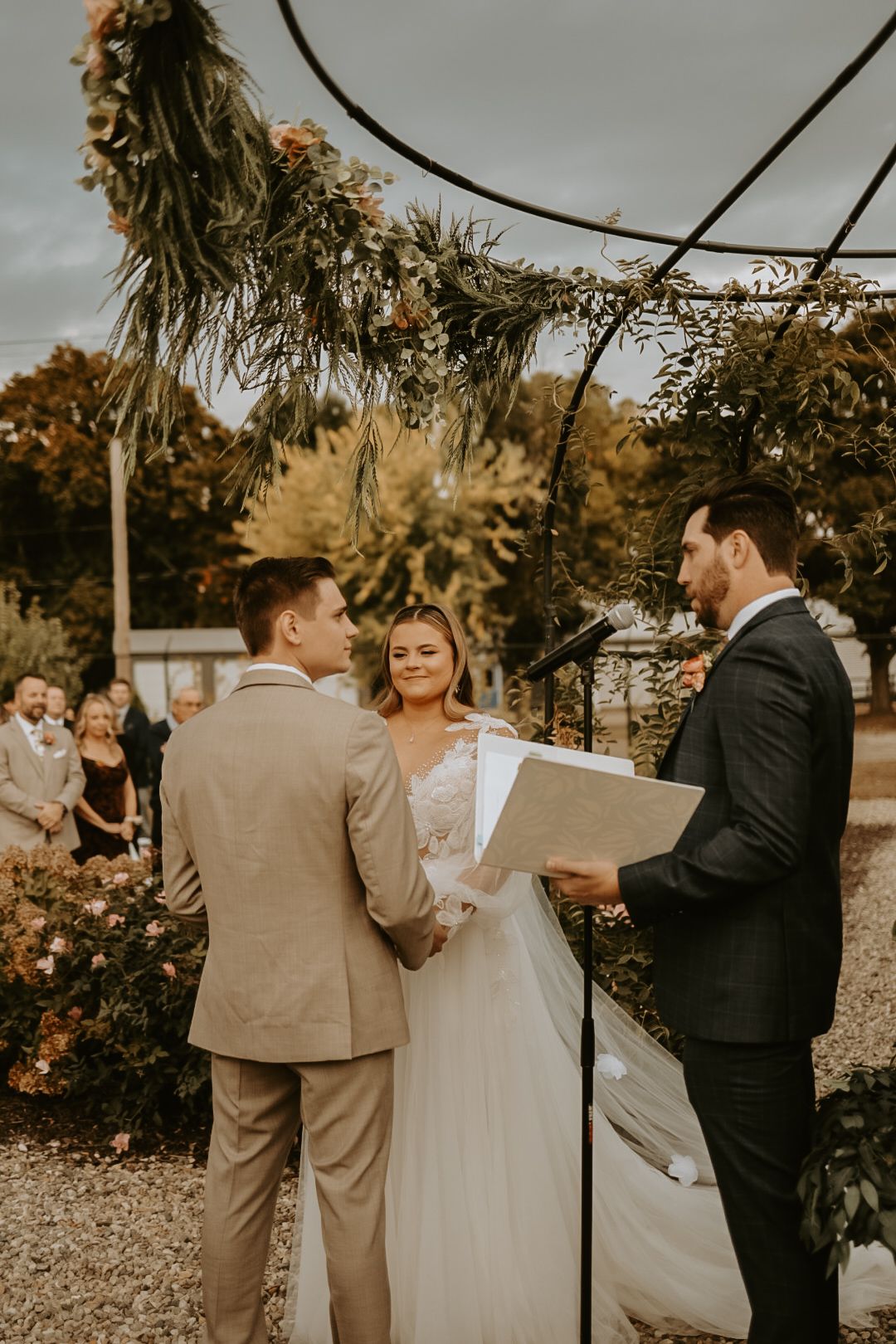 Wedding ceremony in progress under floral arch with officiant and wedding party.