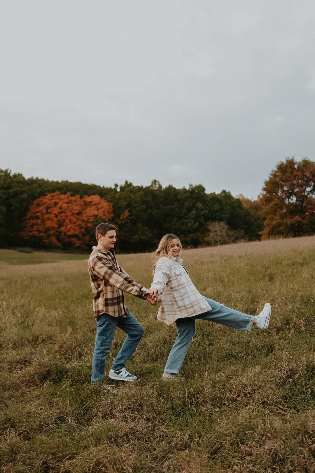 A playful couple wearing plaid shirts and jeans dance together in a golden autumn field.