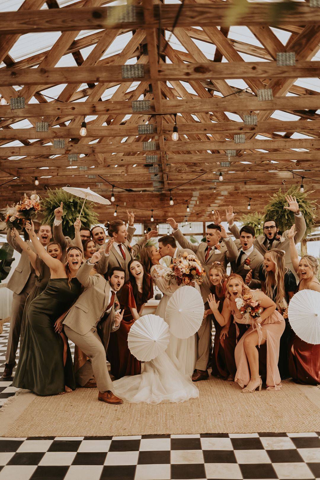 Joyful wedding party celebration under wooden rafters with white parasols and checkered flooring.