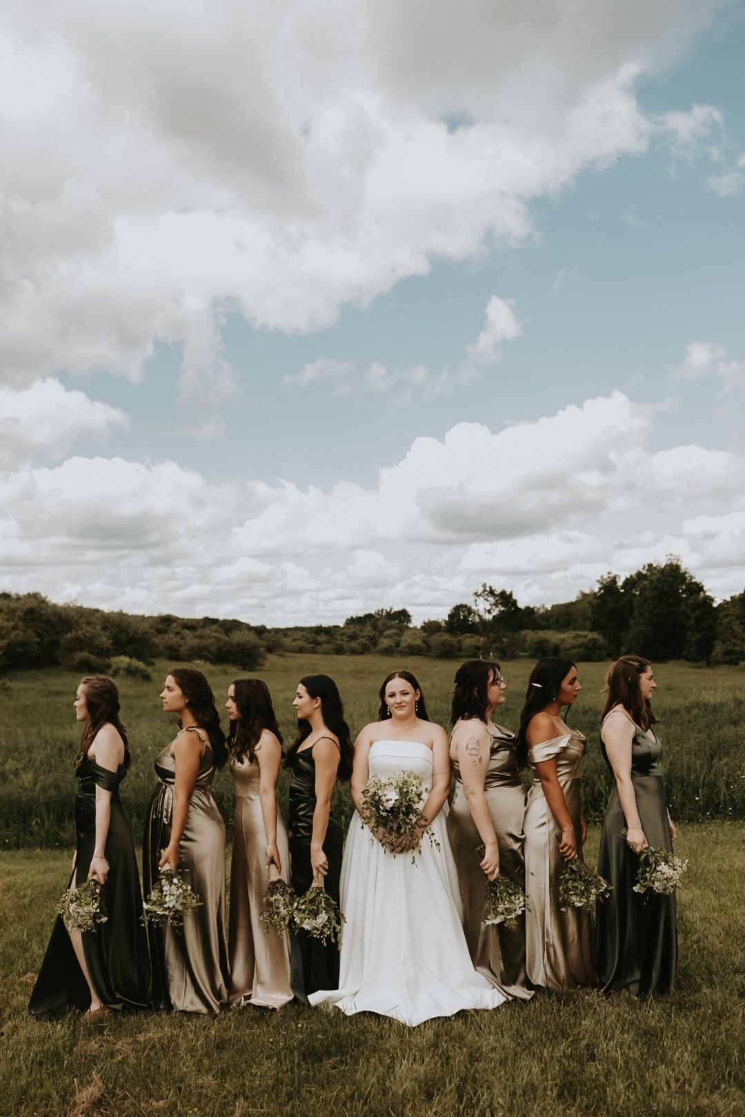 Wedding party in neutral tones poses in a line across a grassy field under cloudy skies.