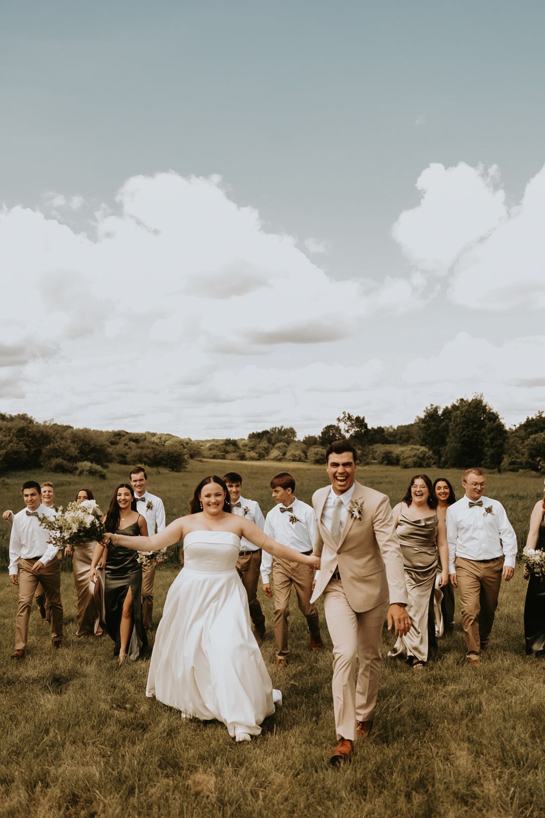 Wedding party in beige and white attire walks together across grassy field.