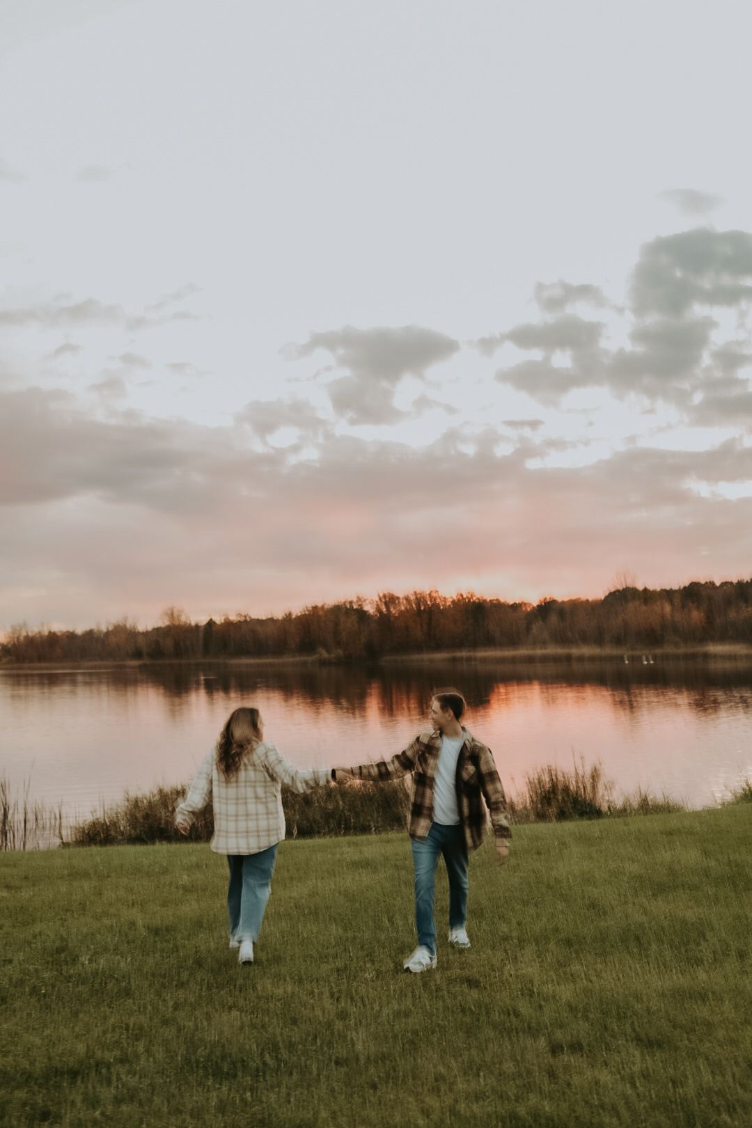 Two people walking on grassy shore with sunset reflecting on calm lake waters.