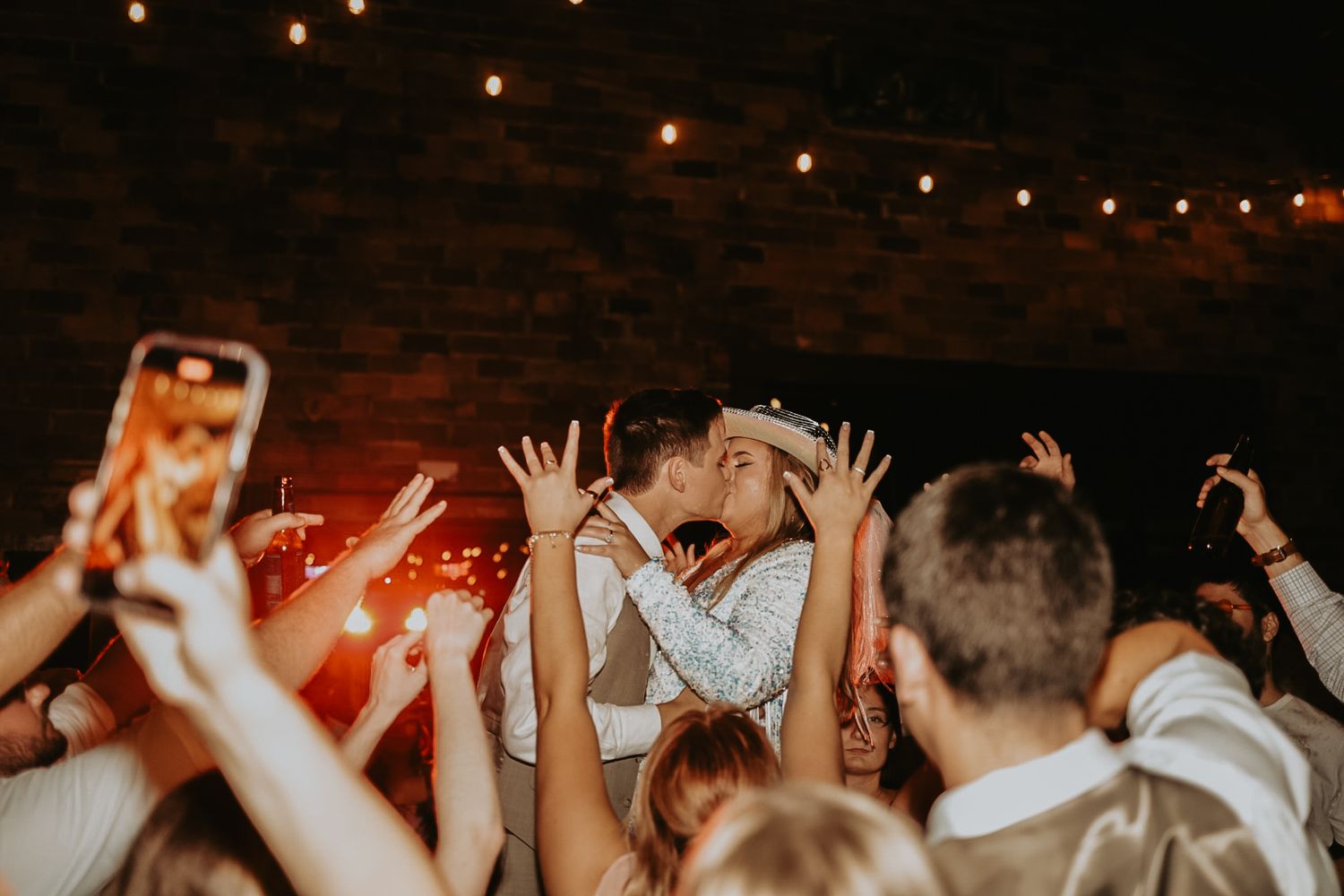 Crowd with raised hands dancing at indoor party under string lights.