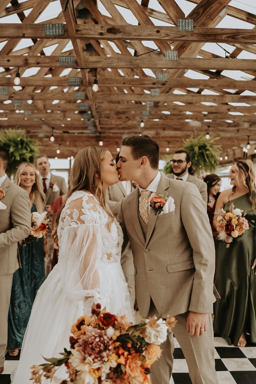 Wedding party members in beige suits and dark dresses celebrate under wooden rafters.