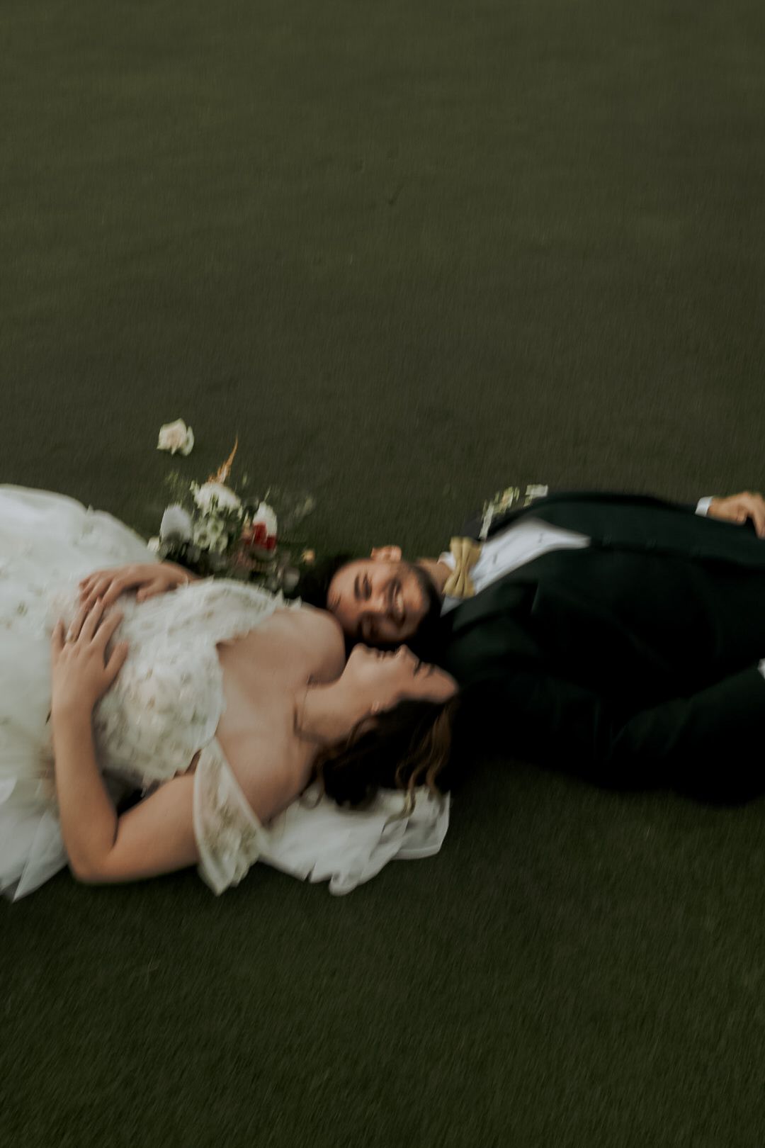A couple in wedding attire lies on grass during a romantic photoshoot.