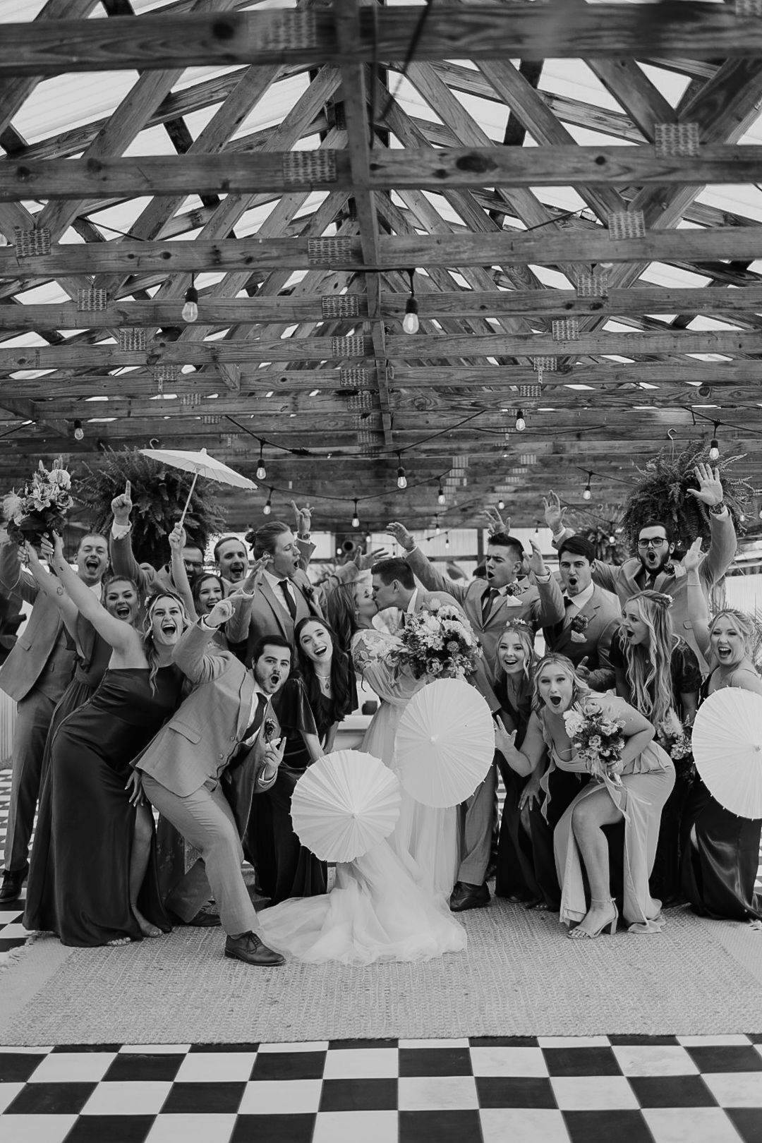 Wedding guests gather for a fun group photo on a checkered dance floor under wooden beams.