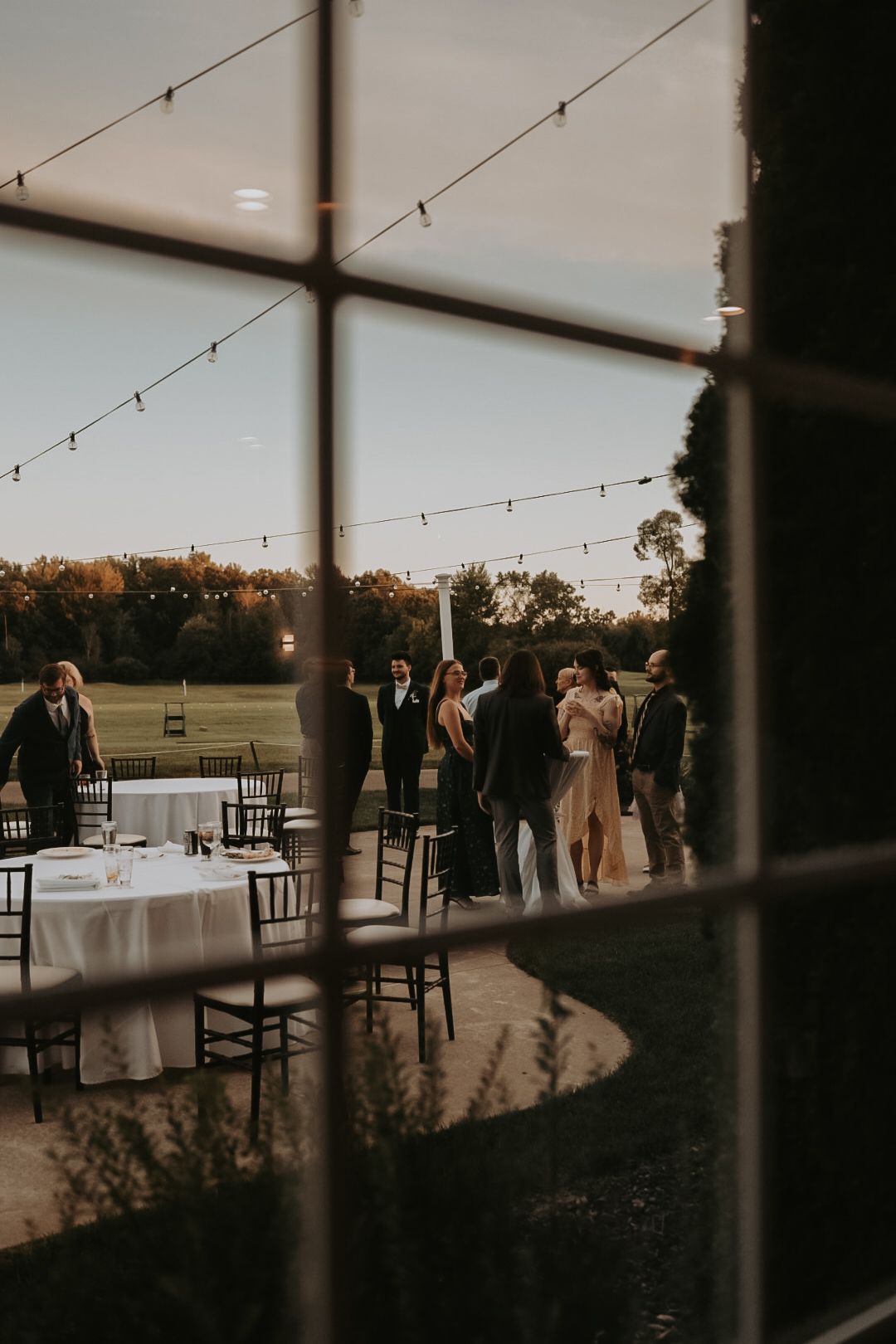 Wedding guests gather on an outdoor patio at sunset with string lights and elegant table settings.