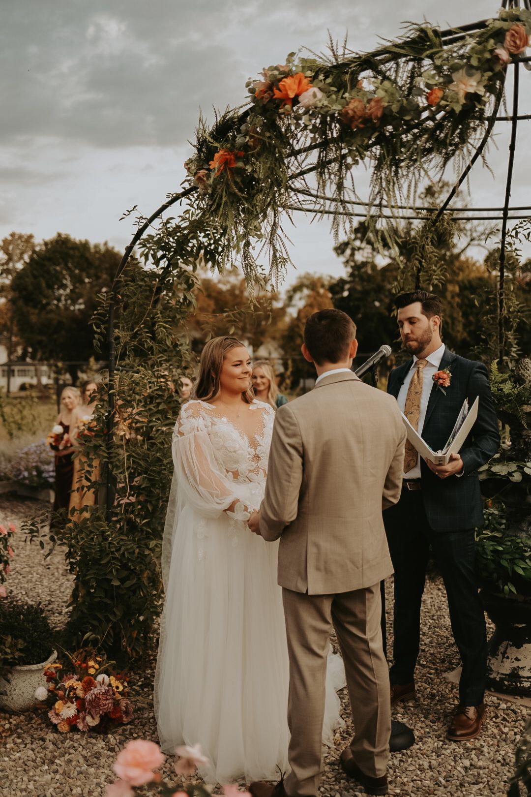 Wedding party gathers under decorated metal gazebo during autumn outdoor ceremony.