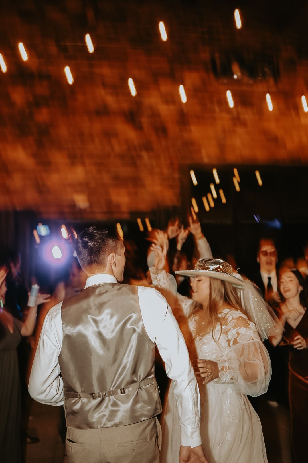 Energetic dance floor scene at a wedding reception with string lights overhead.