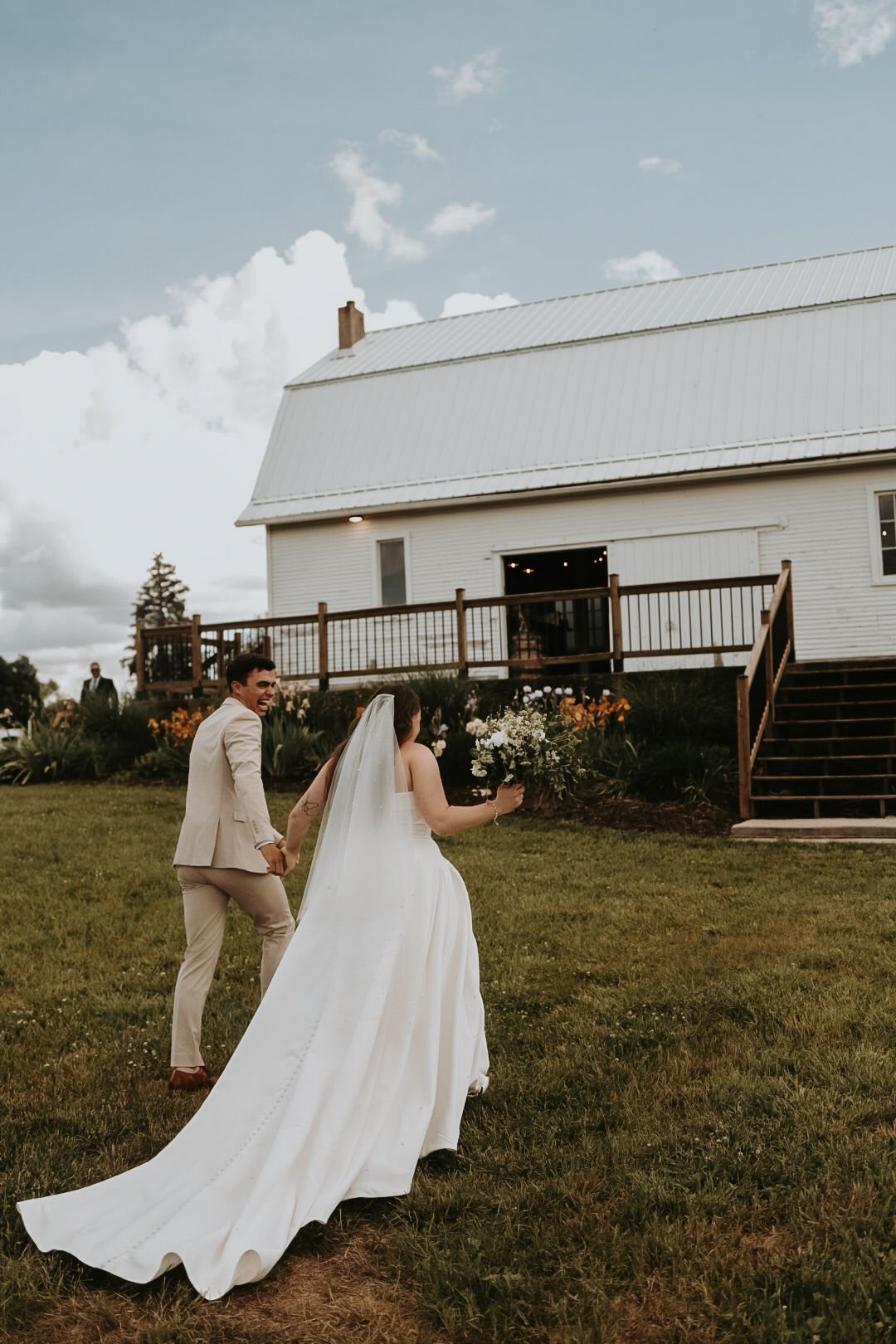 Wedding couple walks near white barn building with flowing dress train.