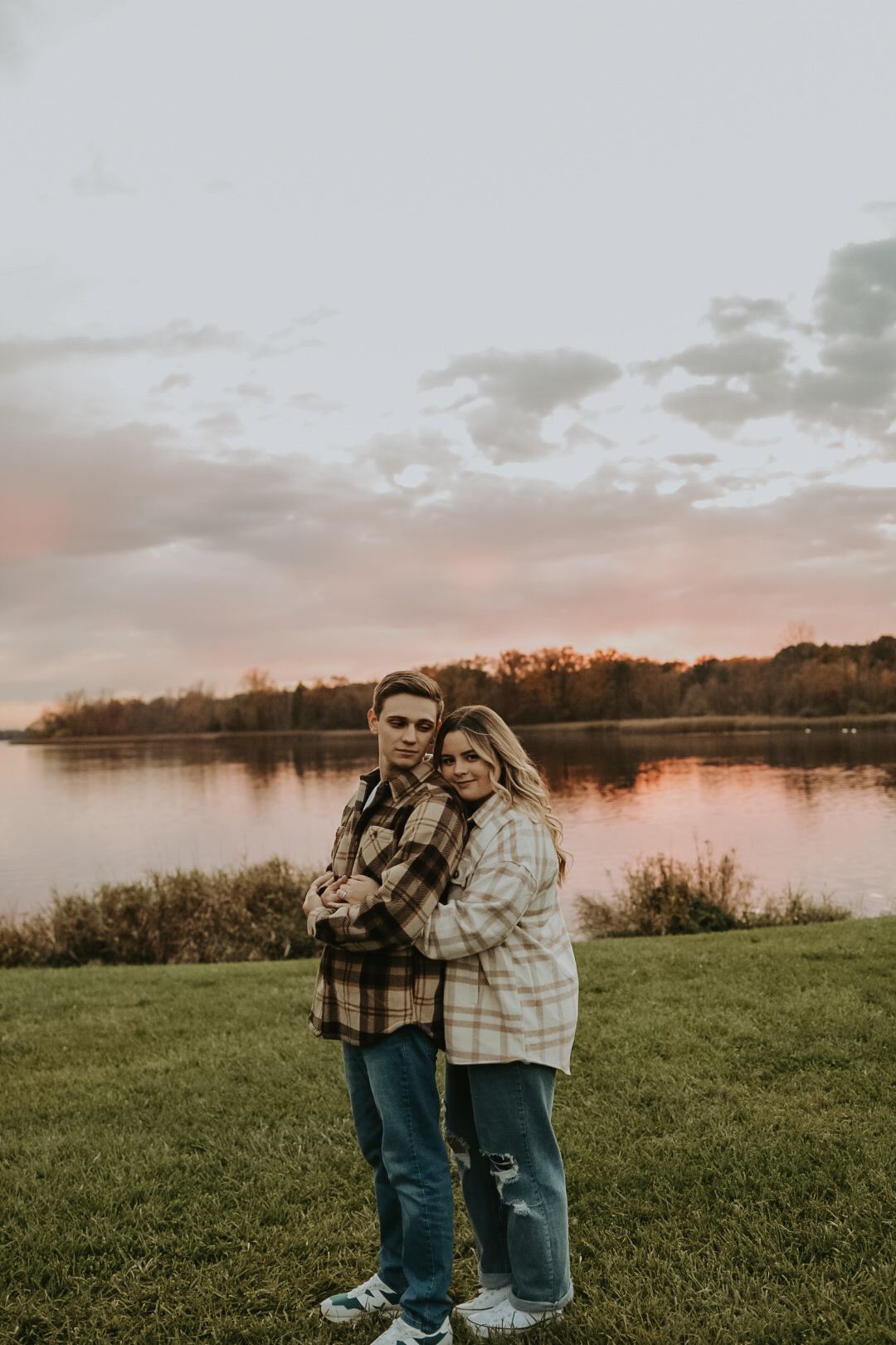 Two people in plaid jackets embrace by a serene lake at dusk with fall foliage in the background.
