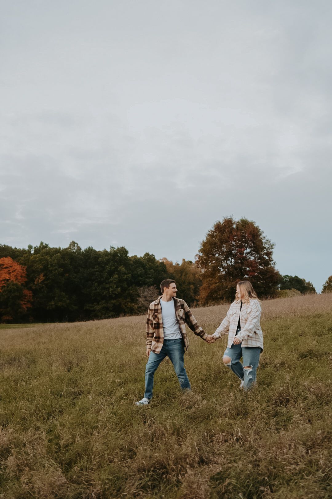 A couple holds hands while walking through a grassy autumn field.